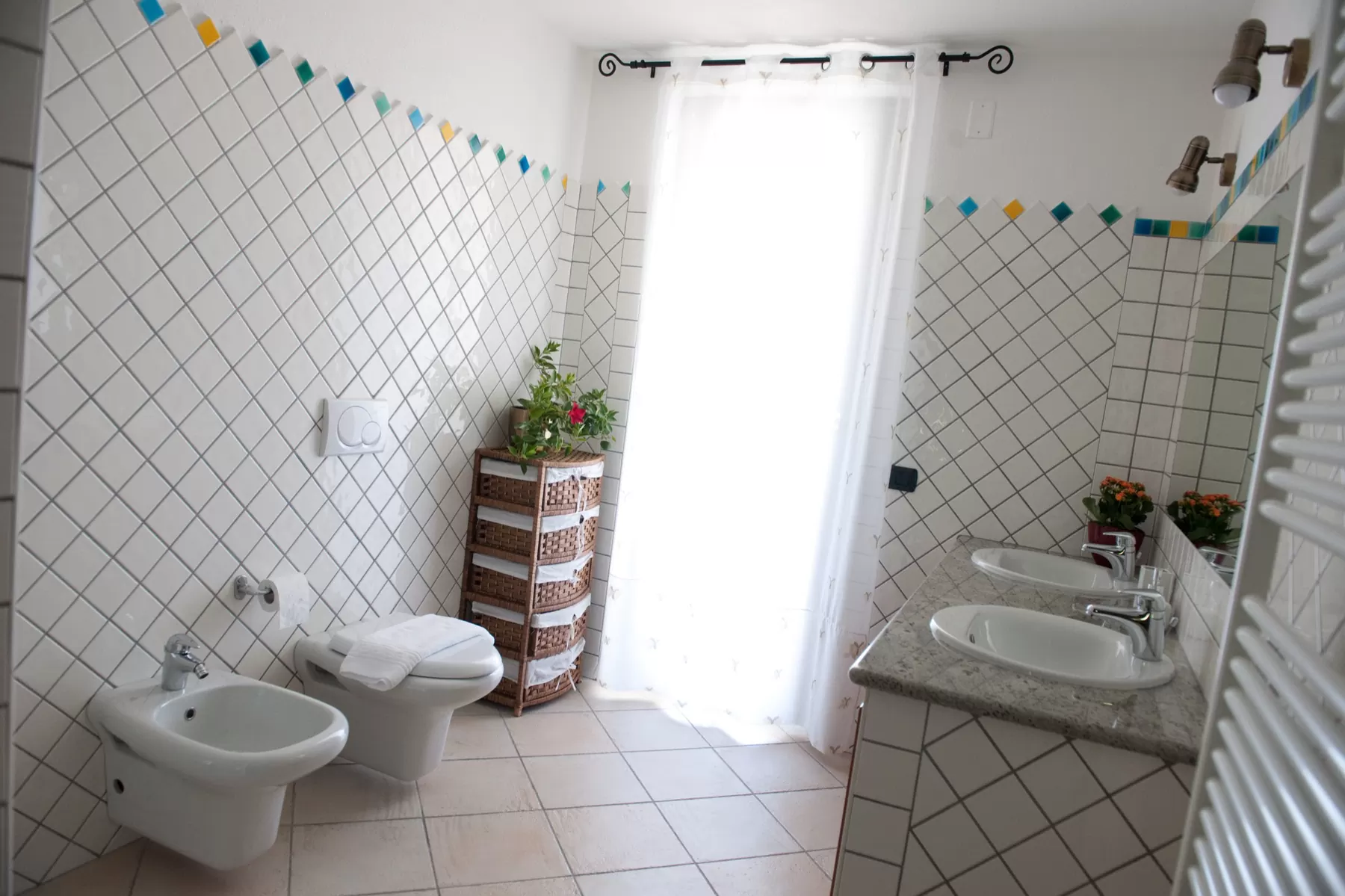 Bagno piastrellato di bianco con doppio lavabo, bidet e una fascia di mattonelle colorate