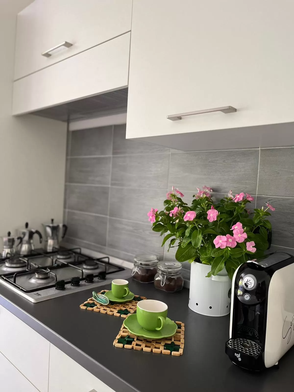Il piano della cucina con le moke sul fornello, due tazzine verdi sui sottopentola e un vaso di fiori rosa