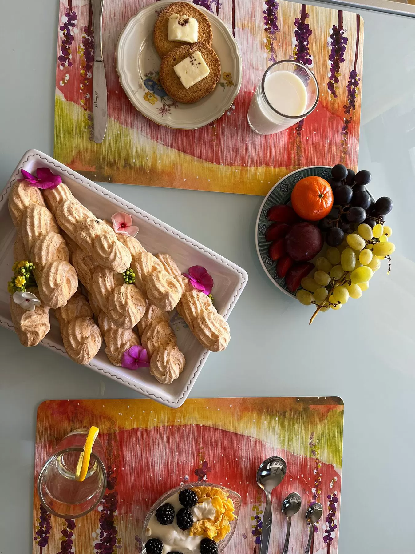 La tavola vista dall'alto: fette biscottate con burro, un bicchiere di latte, il vassoio di dolci con i fiori e un piatto di frutta