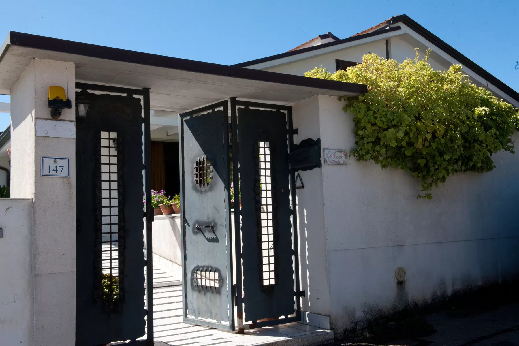 Il cancello in ferro aperto sul vialetto d'ingresso della casa, con l'insegna e la vite sopra il muro