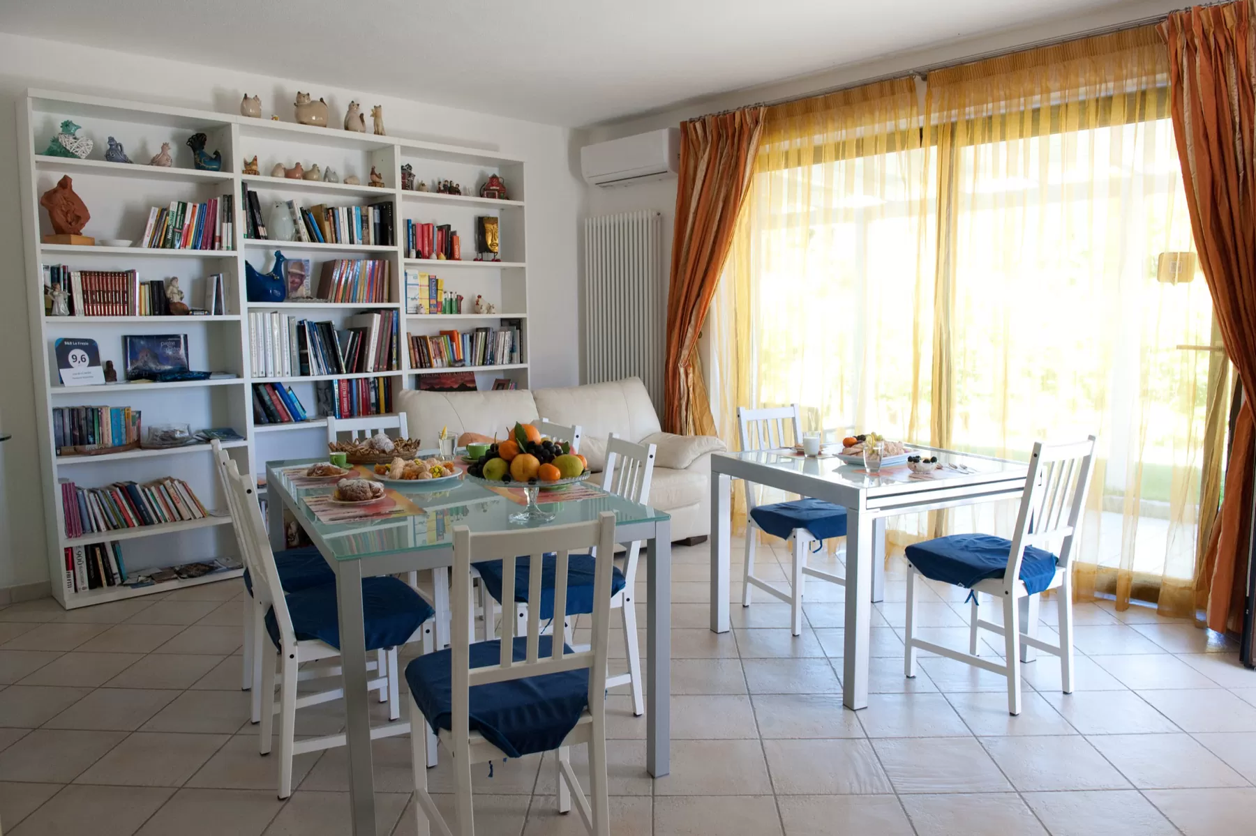 La sala della colazione con la libreria bianca piena di libri e ceramiche, i tavoli apparecchiati e la vetrata con le tende arancioni