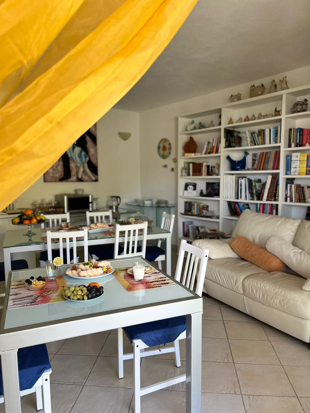 La sala vista da dietro la tenda gialla: il tavolo con i dolci e la frutta, il divano chiaro e la libreria sul fondo